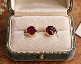 May include: A pair of stud earrings featuring raw, deep red garnet stones set in gold-toned metal. The earrings are displayed side-by-side in a velvet-lined jewelry box with a cream-colored interior and a dark green exterior casing.
