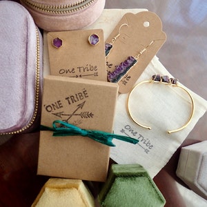Pode incluir: Um conjunto de joias de ametista em uma caixa de presente. O conjunto inclui um par de brincos, uma pulseira e um colar. As joias s&atilde;o embaladas em uma caixa marrom com uma fita verde e as palavras "One Tribe" impressas nela.