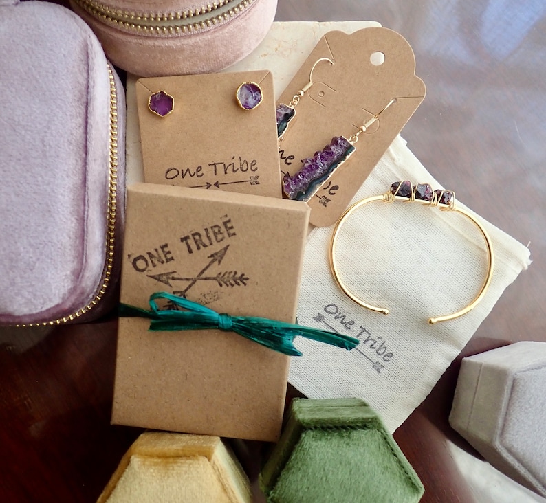 Pode incluir: Um conjunto de joias de ametista em uma caixa de presente. O conjunto inclui um par de brincos, uma pulseira e um colar. As joias s&atilde;o embaladas em uma caixa marrom com uma fita verde e as palavras "One Tribe" impressas nela.