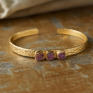 Pode incluir: Uma pulseira tipo bracelete em tom dourado com textura de metal martelado e três pedras preciosas brutas em rosa escuro dispostas em fila no centro. A pulseira tem um design aberto para ajuste e um acabamento polido.