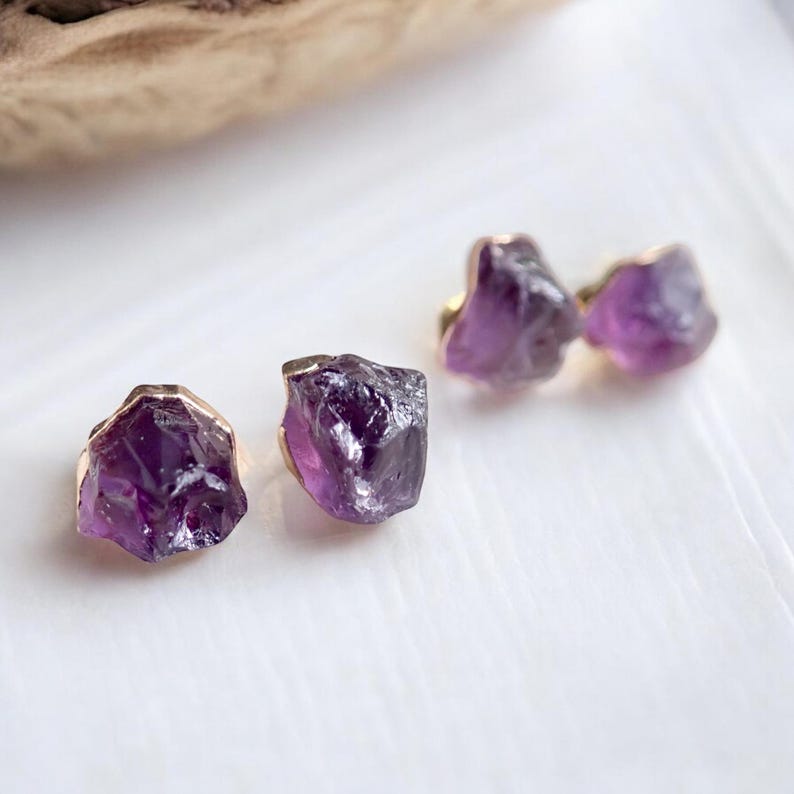 May include: Three raw amethyst stud earrings with gold-colored settings. The earrings are a deep purple color and have a natural, rough texture. The earrings are displayed on a white surface.