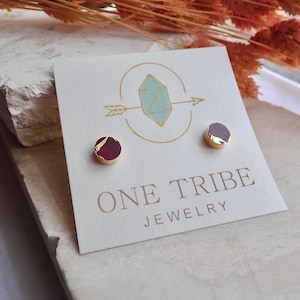 May include: Stud earrings with a gold-coloured border and a dark red stone. The earrings are displayed on a white card with the text "ONE TRIBE JEWELRY" and a logo featuring a crystal and arrow.