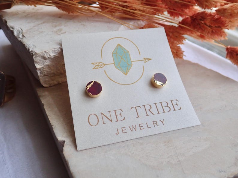 May include: Stud earrings with a gold-coloured border and a dark red stone. The earrings are displayed on a white card with the text "ONE TRIBE JEWELRY" and a logo featuring a crystal and arrow.