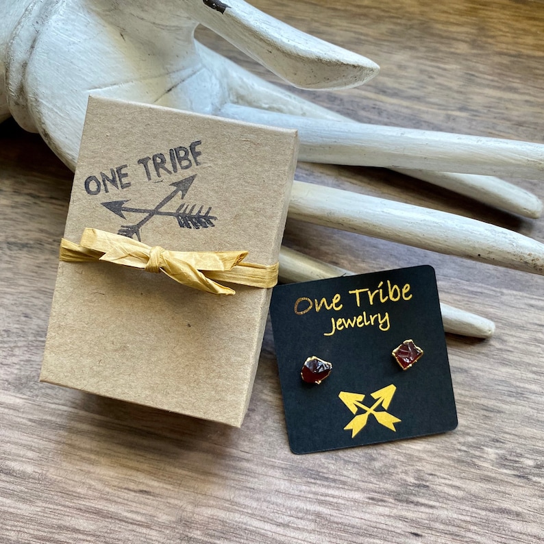 Puede incluir: Un par de pendientes de piedras preciosas rojas con engastes de oro, empaquetados en una peque&ntilde;a caja con las palabras "One Tribe Jewelry" en una tarjeta negra.