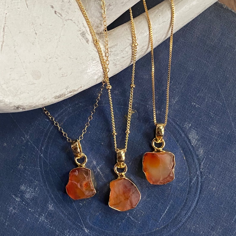 May include: Three gold chain necklaces with carnelian stone pendants. The carnelian stones are all different shapes and sizes. The chains are all different styles.
