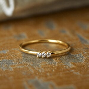 May include: A delicate gold ring featuring three small, clear gemstones set in a row. The ring has a thin band and a polished, shiny finish. The ring is displayed on a textured, brown surface.