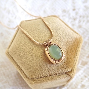 May include: A gold chain necklace with a green oval gemstone pendant. The pendant is set in a gold frame with a decorative edge.