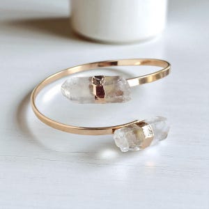 May include: A gold-toned cuff bracelet featuring two clear quartz crystal accents. The crystals are secured with gold-colored metal. The bracelet is displayed on a white surface.
