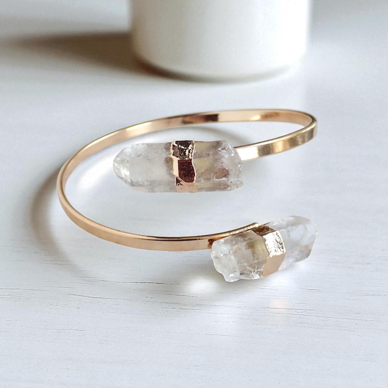 May include: A gold-toned cuff bracelet featuring two clear quartz crystal accents. The crystals are secured with gold-colored metal. The bracelet is displayed on a white surface.