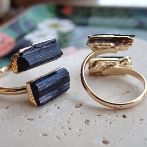 May include: Two gold-colored rings, each featuring a raw, black tourmaline crystal. The crystals are set in a gold-colored metal and are positioned on either side of the ring's band. The rings are displayed on a light-colored surface.