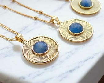 May include: Three gold-colored necklaces with round pendants. Each pendant features a light blue stone set within a sunburst design. The necklaces are displayed on a white marble surface, showcasing the jewelry's elegant design.