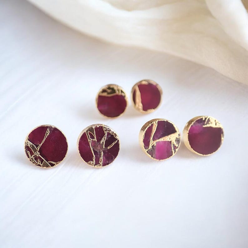 May include: Six pairs of round stud earrings with a gold-coloured trim and a deep red centre. The earrings have a marbled appearance. The earrings are displayed on a white surface.