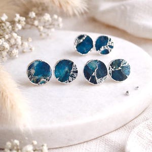May include: A collection of six round stud earrings. Each earring features a dark teal stone with silver-colored metal edging. The earrings are displayed on a white surface with soft, blurred background elements.