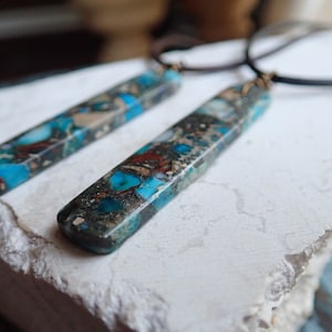 May include: Two long, rectangular pendants made of blue, brown and white marbled stone. The pendants are attached to black leather cords.