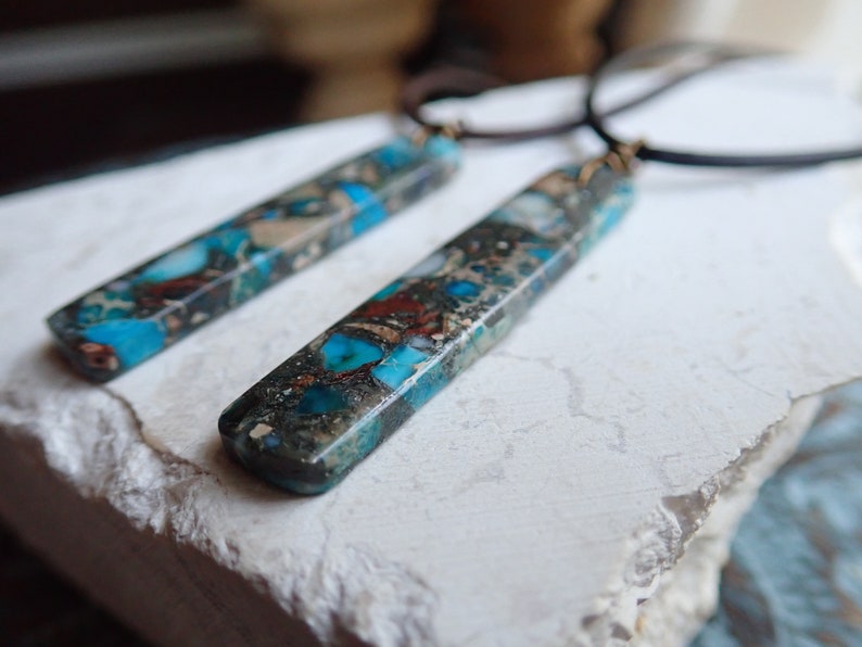 May include: Two long, rectangular pendants made of blue, brown and white marbled stone. The pendants are attached to black leather cords.