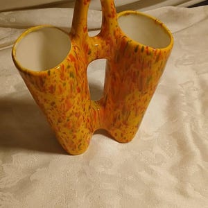 May include: A vintage ceramic vase with a handle, featuring two openings. The vase is painted in a speckled pattern of yellow, orange, and red. The interior of the openings is white.