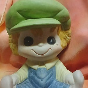 Può includere: Statuetta in ceramica di un ragazzo sorridente con berretto verde, camicia gialla e tuta blu. La statuetta ha capelli biondi, lentiggini e grandi occhi neri. Il ragazzo è seduto con le gambe distese.