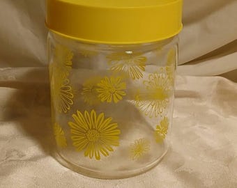 May include: A clear glass jar with a yellow lid, decorated with yellow daisy flower patterns. The jar is cylindrical and appears to be made of glass. The lid is a solid yellow color.