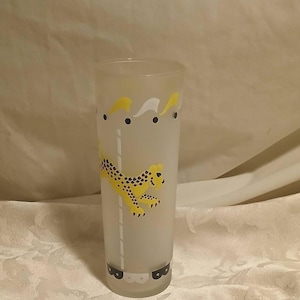 May include: A tall, frosted glass with a yellow and black cheetah design. The glass features a repeating pattern of yellow shapes and black dots. The cheetah is leaping across the glass. A decorative drinking glass.