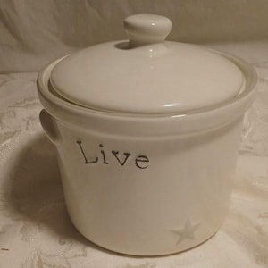 May include: Off-white ceramic jar with a lid and small handles. The word "Live" is printed on the front, along with a star. The jar is designed for kitchen or home decor.