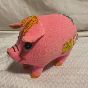May include: A pink stuffed pig with orange accents on its feet and ears. The pig has black stripes on its snout and a tuft of orange yarn for hair. Patches of fabric in various colors are sewn onto its body.