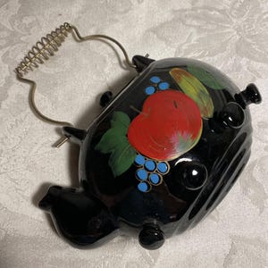May include: Black ceramic teapot with a gold wire handle and decorative fruit design. The teapot features a red apple, blue grapes, and green leaves. The teapot has a unique shape with three small feet.