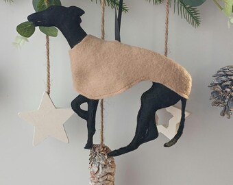 Pode incluir: Um enfeite de feltro de um galgo preto vestindo um suéter bege. O cão está suspenso por barbante, com uma estrela branca e um pinha abaixo. Folhagem e uma pinha no fundo.