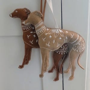 May include: Two felt dog ornaments in shades of brown and tan, with white embroidered details. The dogs are hanging from a white ribbon. The ornaments are likely for home decor or holiday use.