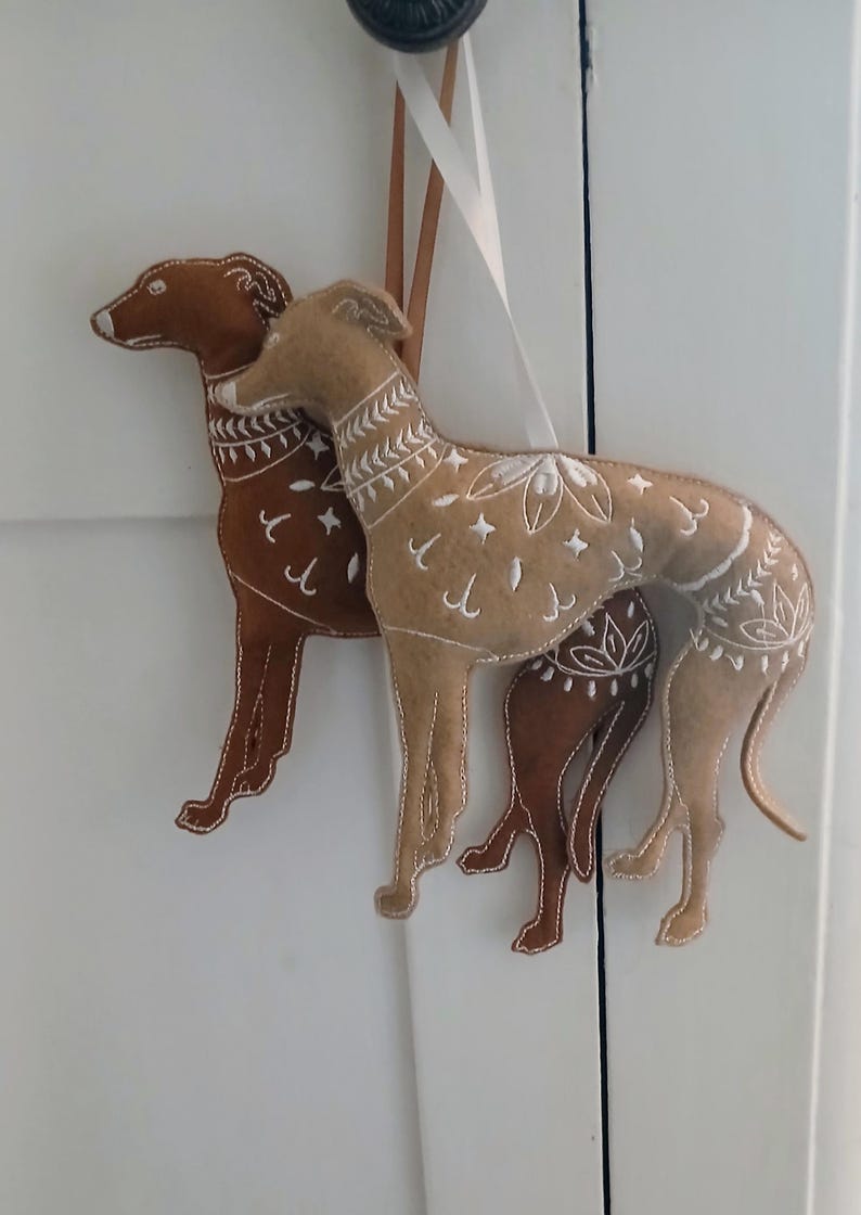 May include: Two felt dog ornaments in shades of brown and tan, with white embroidered details. The dogs are hanging from a white ribbon. The ornaments are likely for home decor or holiday use.