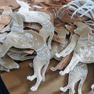 May include: A collection of tan felt dog ornaments with white embroidered details, lying on a wooden surface. The dogs have decorative patterns and are in various poses. Ribbons and scissors are also visible.