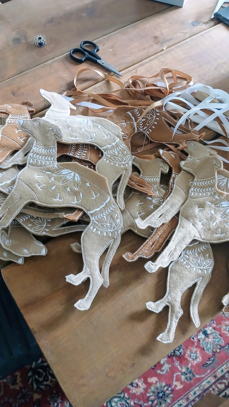 May include: A collection of tan felt dog ornaments with white embroidered details, lying on a wooden surface. The dogs have decorative patterns and are in various poses. Ribbons and scissors are also visible.