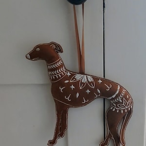 May include: A brown felt dog ornament with white embroidered floral and geometric designs. The dog is hanging from a brown ribbon. The ornament is attached to a white door.