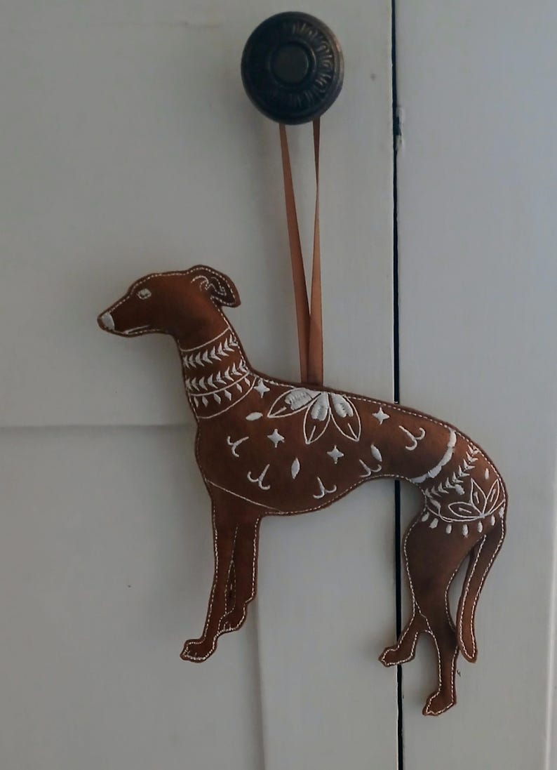 May include: A brown felt dog ornament with white embroidered floral and geometric designs. The dog is hanging from a brown ribbon. The ornament is attached to a white door.