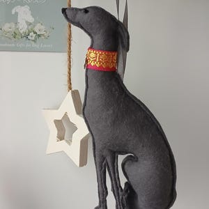 Op de afbeelding: Een grijs vilten hondenornament met een gouden en rode halsband, opgehangen aan een grijs lint. Een witte ster en groen hangen ook. Het ornament is een handgemaakt cadeau voor hondenliefhebbers.