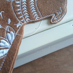 May include: A brown felt dog-shaped applique with white embroidered details. The applique features a detailed design around the neck and head, with a white outline. The dog is partially visible, suggesting it's part of a larger craft project.