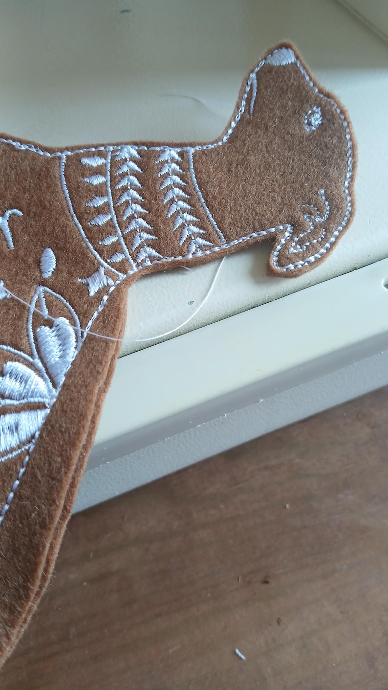 May include: A brown felt dog-shaped applique with white embroidered details. The applique features a detailed design around the neck and head, with a white outline. The dog is partially visible, suggesting it's part of a larger craft project.