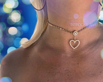 Może przedstawiać: Złoty naszyjnik choker z łańcuszkiem, małym wisiorkiem w kształcie kwiatu i wiszącym wisiorkiem w kształcie serca. Wisiorek w kształcie serca jest ozdobiony małymi, przezroczystymi kamieniami. Na łańcuszku widoczny jest napis "TATIROCKS JEWELRY".