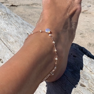 May include: A delicate rose gold anklet with a small, iridescent gemstone and tiny pearls. The anklet is worn on a foot at the beach. The text "TATIROCKS JEWELRY" is visible on the ankle.