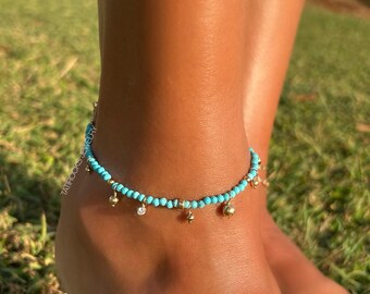 May include: A turquoise beaded anklet with gold-coloured charms and small clear crystals. The anklet is worn on a foot with a grassy background. The anklet is from TATROCKS JEWELRY.