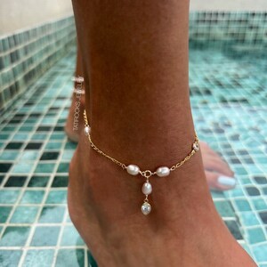 以下が含まれることがあります： 小さなパールとクリスタルアクセントが施された、繊細なゴールドチェーンのアンクレット。アンクレットには、パールとクリスタルチャームがぶら下がっています。