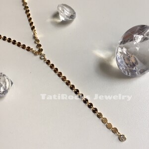 May include: A gold chain necklace with small round discs and two clear crystal accents. The necklace is on a white surface with two clear crystal shapes.