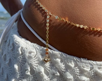 May include: Gold body chain with a heart charm. The chain is made of small, round gold links and is worn around the waist. The heart charm is attached to the chain by a small gold loop.