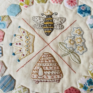 Könnte beinhalten: Gestickte Textilkunst mit einer Biene mit Krone, einer Krone, einem Bienenstock und Blumen, alles mit gelbem, braunem und blauem Faden auf cremefarbenem Hintergrund gestickt. Das Design wird von bunten, gemusterten Stoffsechsecken umrahmt.