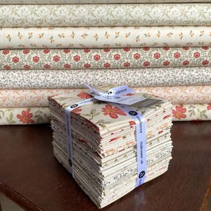 Könnte beinhalten: Ein Stapel Stoffquadrate, gebunden mit einem weißen Band, mit floralen Mustern in Rot, Creme und Grün. Der Hintergrund zeigt Stoffrollen mit ähnlichen floralen Designs, ideal für Quilt- und Bastelprojekte.
