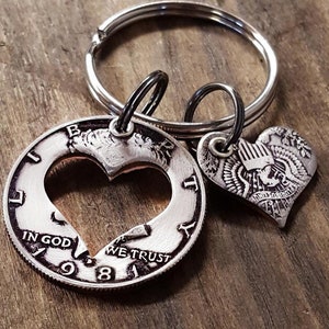May include: A silver-colored keychain featuring a heart-shaped cutout coin from 1984, with the words "IN GOD WE TRUST." A smaller heart-shaped charm is also attached. The keychain is on a silver ring.