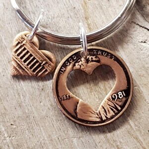 May include: A copper-colored keychain with two charms. One charm is a heart-shaped coin with a heart cut out of the center, the year 1981, and the words "In God We Trust" and "Liberty". The other charm is a heart-shaped design.