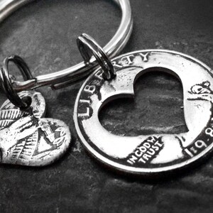 May include: Silver-toned keychain featuring a heart-shaped charm and a coin with a heart cut-out. The coin displays the words "LIBERTY" and "IN GOD WE TRUST" along with the year "1981". The keychain is attached to a silver ring.