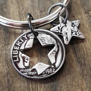 May include: A silver-colored keychain featuring a cut-out coin with a star-shaped design and a smaller star charm. The coin is dated 1982 and has the words "LIBERTY" and "IN GOD WE TRUST" engraved on it. The keychain is on a wooden surface.