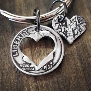 May include: A silver-colored keychain featuring two heart-shaped charms. One charm is a coin with a heart cut out, the word "LIBERTY" is visible. The other charm is a smaller heart-shaped coin. The year "1982" is visible on the larger coin.