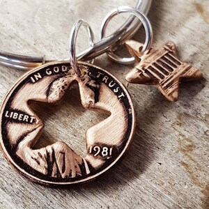 May include: A copper-colored keychain featuring a cut-out star design on a 1981 penny, with a small star charm. The penny has the words "IN GOD WE TRUST" and "LIBERTY" engraved on it. The keychain is attached to a silver ring.